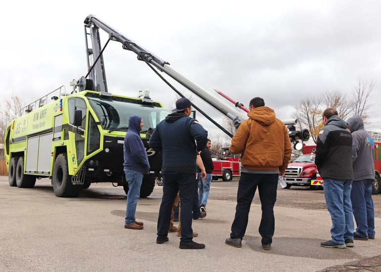 A Striker 6x6 ARFF with the Snozzle extended outside on a cloudy day with men standing nearby.